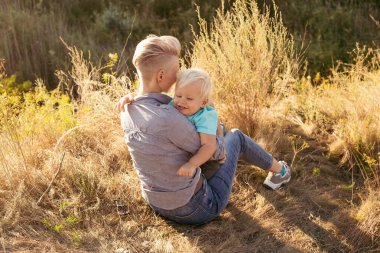 Genç mutlu anne yazın doğada oğlunu kucaklıyor çimlerin üzerinde oturuyor gün batımında yeşil tepelerin yakınında güneş ışınlarıyla.