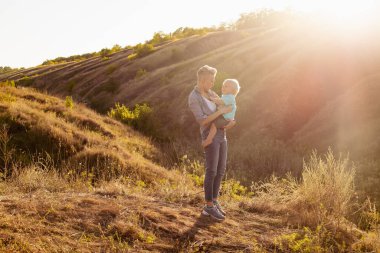Anne ve oğlu yazın doğada vakit geçiriyorlar. Güneş ışınları günbatımında yeşil tepelerin yakınında.