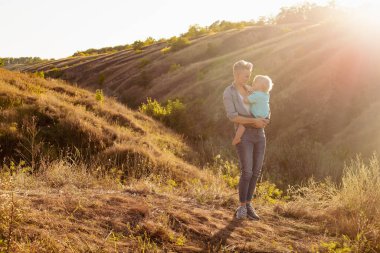 Anne ve küçük oğlu yazın güneşin batışında yeşil tepelerin yakınında güneşin tadını çıkarıyorlar.