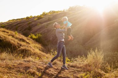 Anne ve küçük oğlu oynuyorlar ve yazın gün batımında yeşil tepelerin yakınında eğleniyorlar. Anne bebeği kaldırıyor.