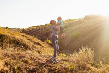 Annem küçük oğlunu yazın doğada havaya fırlatır güneş ışınlarıyla birlikte yeşil tepelerin yakınındaki çimlere.