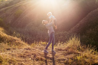 Anne, yaz mevsiminde küçük oğlunu çimlerin üzerinde, günbatımında yeşil tepelerin yakınında, elleriyle havaya kaldırır.
