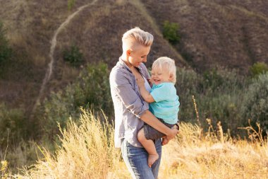 Anne, yaz aylarında doğada oynarken oğlunu kucaklıyor. Gün batımında yeşil tepelerin yakınında güneş ışınlarıyla oynuyor.