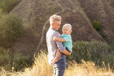 Mutlu gülümseyen genç anne, yaz mevsiminde doğada oynarken oğlunu kucaklıyor. Gün batımında yeşil tepelerin yakınında güneş ışınlarıyla oynuyor.