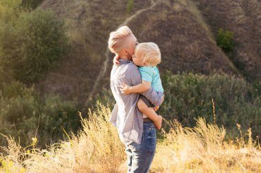 Mutlu gülümseyen genç anne, yaz mevsiminde doğada oynarken oğlunu kucaklıyor. Gün batımında yeşil tepelerin yakınında güneş ışınlarıyla oynuyor.