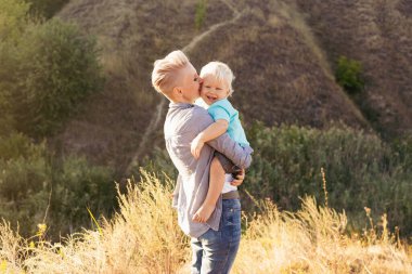 Mutlu anne oğlunu doğada kollarında tutuyor. Güneş ışınları günbatımında yeşil tepelerin yakınında.
