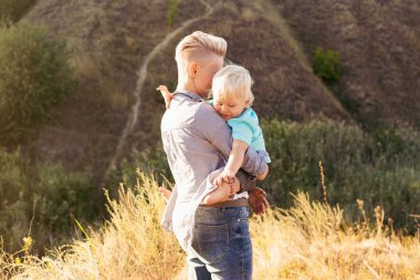 Mutlu anne oğlunu doğada kollarında tutuyor. Güneş ışınları günbatımında yeşil tepelerin yakınında.