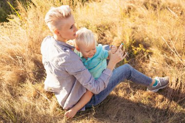 Genç mutlu anne yazın doğada oğlunu kucaklıyor çimlerin üzerinde oturuyor gün batımında yeşil tepelerin yakınında güneş ışınlarıyla.