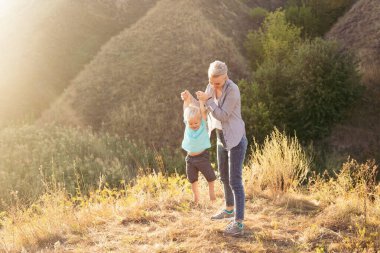 Anne yaz aylarında doğada oynarken oğlunu kaldırır. Güneş ışınları günbatımında yeşil tepelerin yakınında.