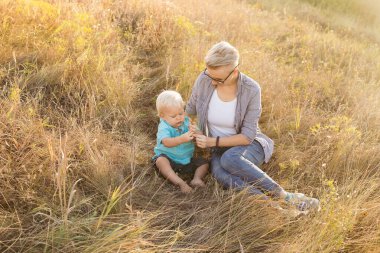 Oğlan ve annesi yazın birlikte doğada vakit geçiriyorlar gün batımında güneş ışınlarıyla çimlerin üzerinde oturuyorlar.