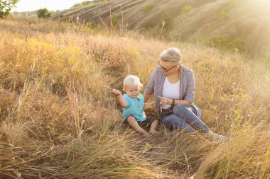 Anne ve küçük oğlu yazın birlikte doğada vakit geçiriyorlar gün batımında güneş ışınlarıyla çimenlerin üzerinde oturuyorlar.