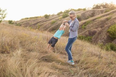 Anne ve küçük oğlu oynuyorlar ve yazın gün batımında yeşil tepelerin yakınında eğleniyorlar. Anne bebeği kaldırıyor.