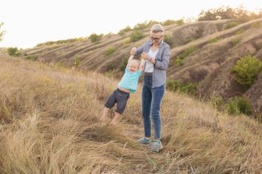 Anne ve küçük oğlu oynuyorlar ve yazın gün batımında yeşil tepelerin yakınında eğleniyorlar. Anne bebeği kaldırıyor.