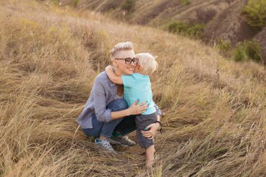 Anne, yazın doğada oğlunu kucaklıyor çimlerin üzerinde oturuyor gün batımında yeşil tepelerin yakınında güneş ışınlarıyla.