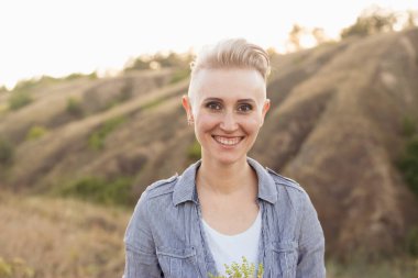 Saçları kısa kesimli, gülümseyen bir kadın. Doğada mavi gömlek giyiyor. Gün batımında yeşil tepelerin yakınında.