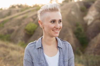 Kısa saçlı, mutlu bir kadın. Doğada açık havada mavi gömlek giyiyor. Gün batımında yeşil tepelerin yakınında.