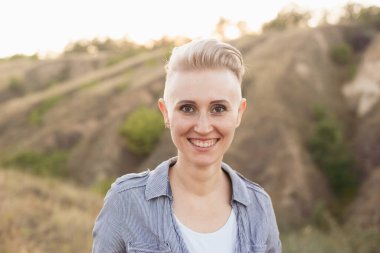 Kısa saçlı, mutlu genç bir kadın. Doğada mavi gömlek giyiyor. Gün batımında yeşil tepelerin yakınında.