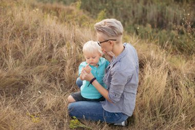 Anne, yazın doğada oğlunu kucaklıyor çimlerin üzerinde oturuyor gün batımında yeşil tepelerin yakınında güneş ışınlarıyla.