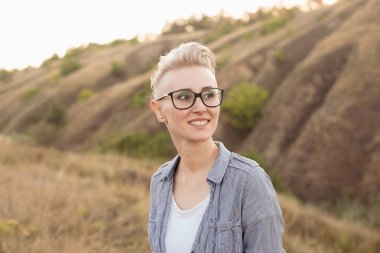 Kısa saçlı, kısa saçlı, gözlüklü, açık havada mavi gömlekli, günbatımında yeşil tepelerin yakınında şık bir kadın.