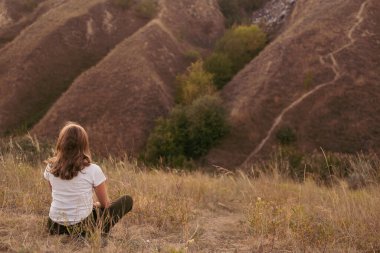 Doğada çimlerin üzerinde oturan kadının arka planı. Gün batımında yeşil tepelerin yakınında.