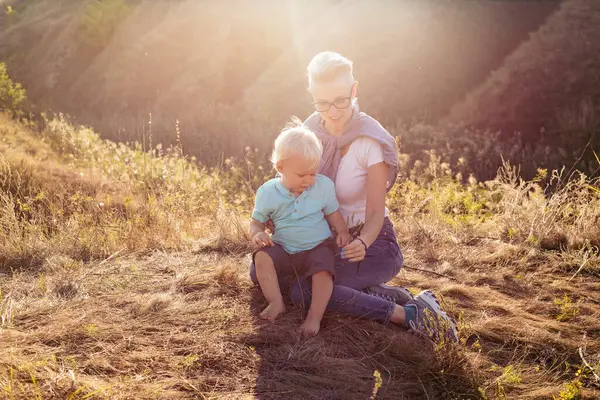 Yazın doğada, gün batımında güneş ışığıyla çimlerin üzerinde oturan bebek ve annesi.