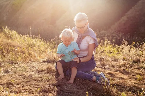 Yazın doğada, gün batımında güneş ışığıyla çimlerin üzerinde oturan bebek ve annesi.