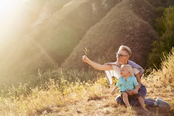 Anne, küçük oğluyla yazın doğada oynuyor çimlerin üzerinde oturuyor gün batımında yeşil tepelerin yakınında güneş ışınlarıyla.