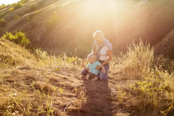 Anne, küçük oğluyla yazın doğada oynuyor çimlerin üzerinde oturuyor gün batımında yeşil tepelerin yakınında güneş ışınlarıyla.