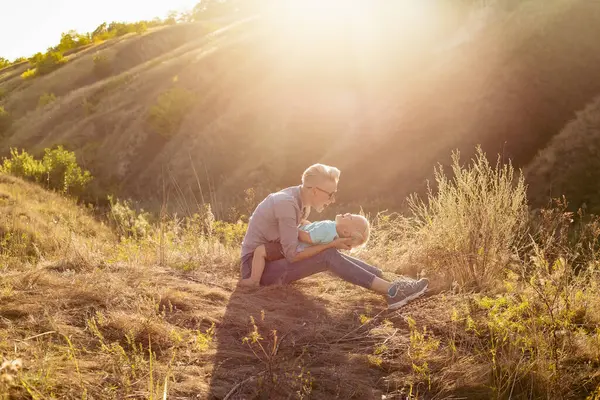 Anne, küçük oğlunu yazın doğada kucaklıyor çimlerin üzerinde oturuyor gün batımında yeşil tepelerin yakınında güneş ışınlarıyla.
