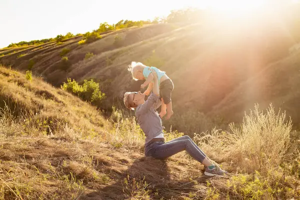 Anne, yazları doğada oğlunu kaldırıyor çimlerin üzerinde oturuyor gün batımında yeşil tepelerin yakınında güneş ışınlarıyla.