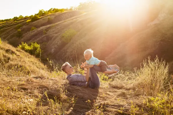 Anne gülümsüyor ve oğlunu doğada doğada kaldırıyor. Yeşil tepelerin yakınında güneş ışınlarıyla çimlerin üzerinde oturuyor.