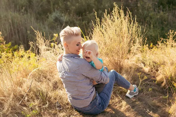 Anne gülüyor ve oğlunu kucaklıyor yazın çimlerin üzerinde oturuyor gün batımında yeşil tepelerin yakınında güneş ışınlarıyla