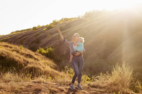 Anne doğada küçük bebeğiyle, anne çocuğa bir şey gösteriyor, gün batımında güneş ışınları.