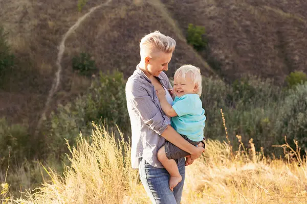 Anne, yaz aylarında doğada oynarken oğlunu kucaklıyor. Gün batımında yeşil tepelerin yakınında güneş ışınlarıyla oynuyor.
