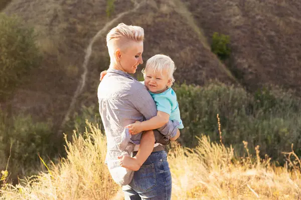 Mutlu anne oğlunu doğada kollarında tutuyor. Güneş ışınları günbatımında yeşil tepelerin yakınında.