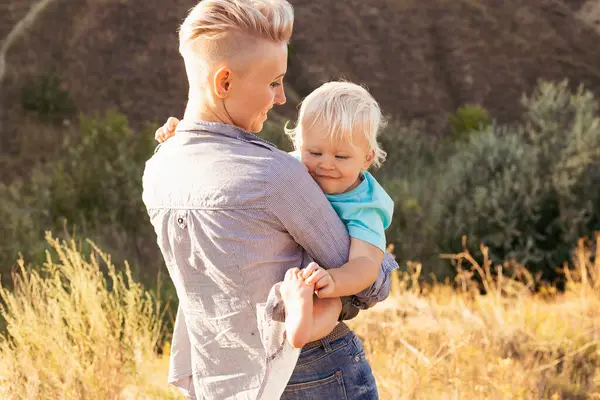 Genç, mutlu bir anne yazın doğada oğlunu kucaklıyor. Gün batımında yeşil tepelerin yakınında güneş ışınlarıyla.