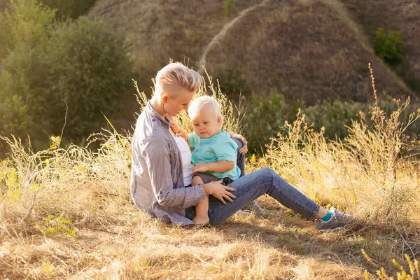 Genç mutlu anne yazın doğada oğlunu kucaklıyor çimlerin üzerinde oturuyor gün batımında yeşil tepelerin yakınında güneş ışınlarıyla.