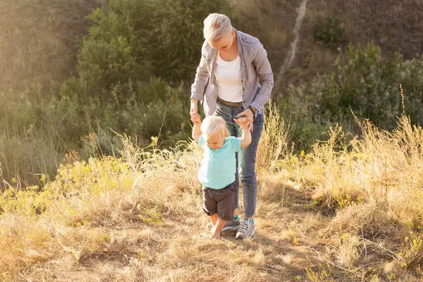 Küçük çocuk ilk adımlarını atıyor ve anne yazın ellerini çimlerin üzerinde gün batımında güneş ışınlarıyla tutuyor.