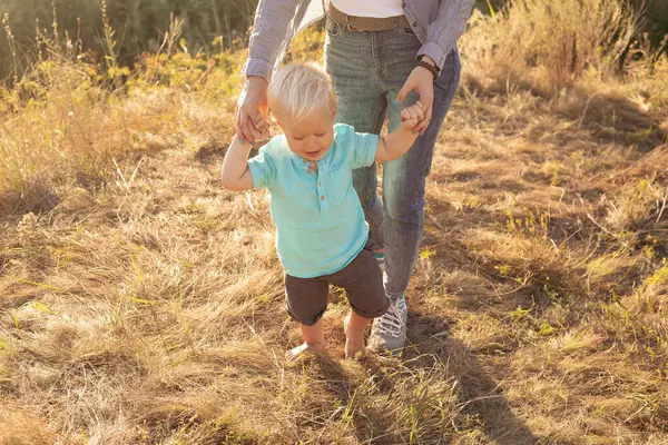 Küçük çocuk ilk adımlarını atıyor ve anne yazın ellerini çimlerin üzerinde gün batımında güneş ışınlarıyla tutuyor.