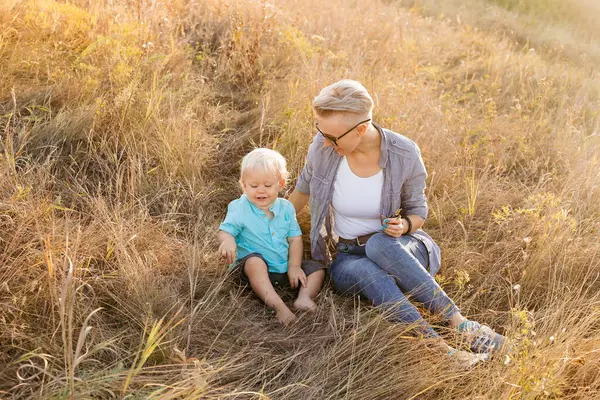 stock image Baby boy and his mother in nature in summer sitting on grass with sun rays on sunset