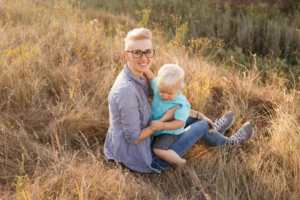 Mutlu anne yazın doğada oğlunu kucaklıyor çimlerin üzerinde oturuyor gün batımında yeşil tepelerin yakınında güneş ışınlarıyla.