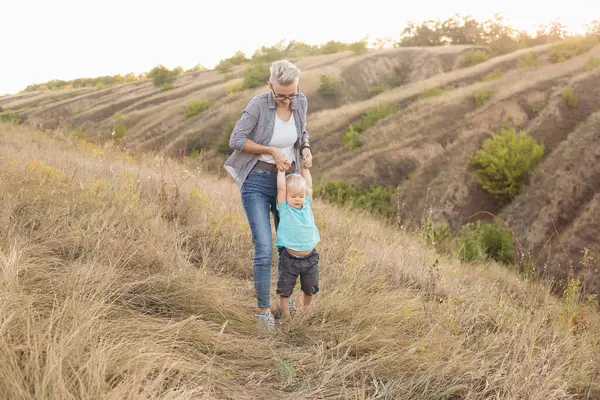 Küçük erkek çocuk ilk adımlarını atıyor ve anne yazın ellerini çimlerin üzerinde gün batımında güneş ışınlarıyla tutuyor.