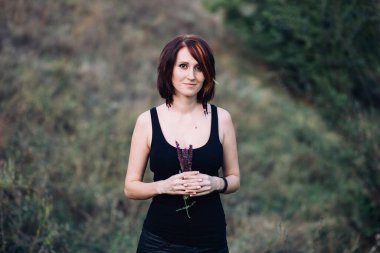 Doğada, günbatımında yeşil tepelerin yakınında elinde çiçeklerle gezen Boho hippi kadın.