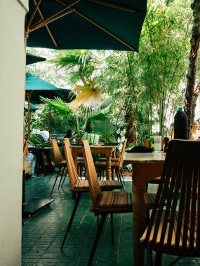 Görüntü, yemyeşil bitkilerle çevrili ahşap sandalyeleri ve masaları olan ve büyük şemsiyelerle gölgelenmiş, tropikal bir ortam sunan sakin bir açık kafe yemek alanını gösteriyor..