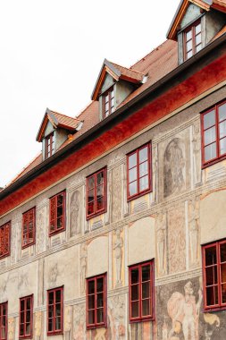 A beautifully preserved historic building features intricate murals depicting various figures and scenes, complemented by striking red framed windows. The architecture reflects the charm of a European town, showcasing artistic details and vibrant col clipart