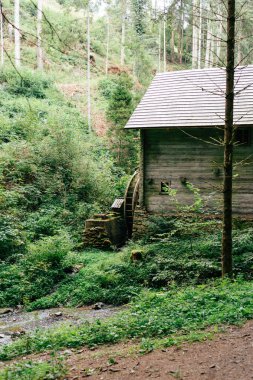 The watermill stands surrounded by vibrant greenery, with trees towering in the background and a gentle stream trickling by, creating a peaceful atmosphere. clipart