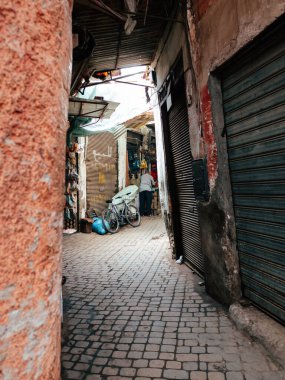 Dar bir sokak, çeşitli mallarla dolu canlı bir pazara açılıyor. Duraklar yol boyunca uzanır ve bir bisiklet duvara yaslanır, canlı atmosferi artırır..