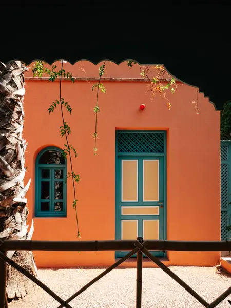 stock image An orange structure with blue doors and windows stands amidst greenery, showcasing unique architectural details and tranquility in a well-lit courtyard.