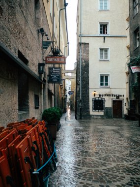 Dar, kaldırım taşlı bir ara sokak son yağmuru yansıtıyor. Gri gökyüzünün altındaki kapalı restoranların önünde istiflenmiş sandalyelerle huzurlu bir atmosfer sergiliyor..