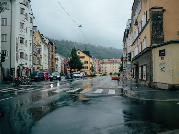 stock image A serene street drenched in rain shows glistening pavements and a few cars. Colorful buildings line the road, with misty mountains visible in the distance.